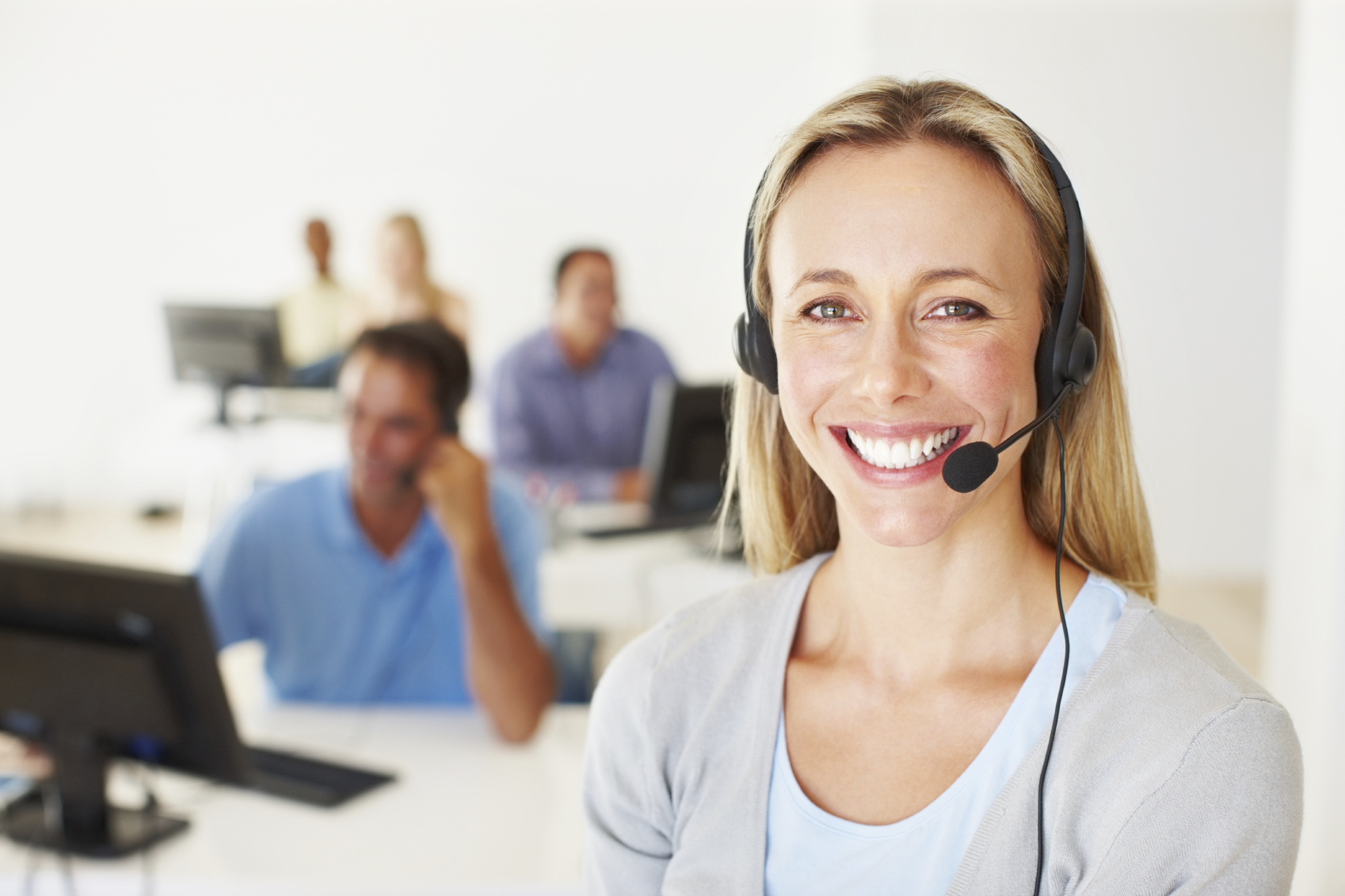 An image of a call center agent smiling with her headset on. There are also various team members behind her taking calls at their desks.